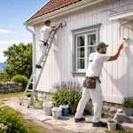 Fasadmålning av trähus på Gotland i kustnära miljö – två målare i arbete mot havsutsikt med penslar och färgburkar. Filnamn: fasadmalning-gotland-trahus-kustmiljo